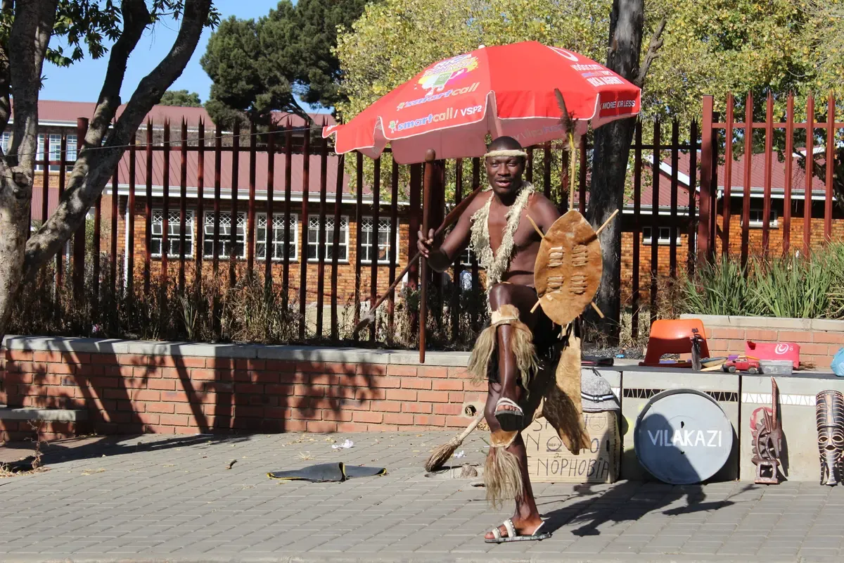 A street performer working the pavement of Vilakazi Street in Soweto, trees and houses behind